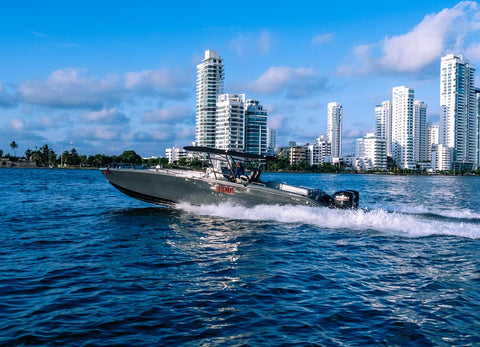A gray motorboat with a hardtop and T-top sail structure speeds across blue water, leaving a white wake, with a backdrop of white high-rise buildings and a blue sky with clouds.