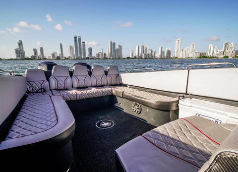 The cockpit seating area of a luxury motorboat with quilted gray and white upholstery, set against a backdrop of water and a city skyline under a blue sky.