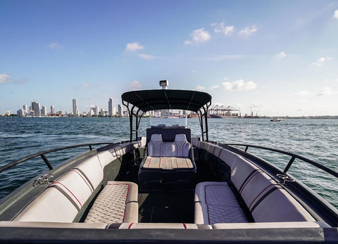 A luxury motorboat with cream and gray upholstered seating and a black hardtop canopy, photographed from the rear looking forward on open water with a distant city skyline under a blue sky.