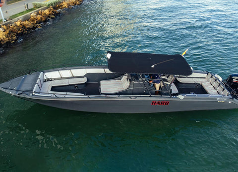 A gray aluminum boat with a black bimini top canopy, gray and white seating, and a black outboard motor, shown floating on greenish-blue water. The side of the boat features the red 'HARB' brand name.