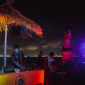 DJ at a tropical outdoor event with colorful lighting and a thatched roof.