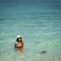 Woman in a bikini and hat standing in clear ocean water with a dog nearby.
