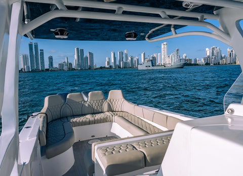 A luxury boat interior with cream and gray tufted seating, photographed from the open deck looking out toward a blue ocean and a city skyline in the distance.